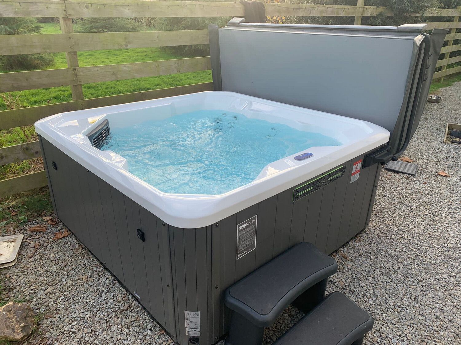 Hot tub with blue water, gray exterior, and open cover, sitting on gravel next to a wooden fence.