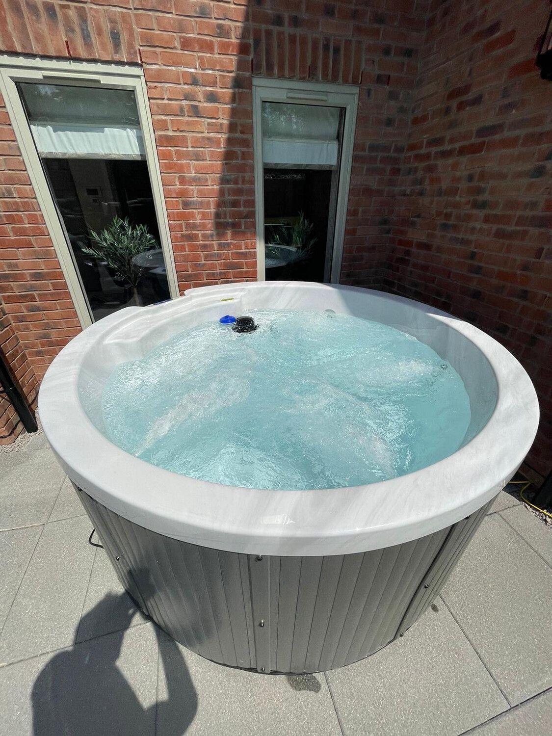 A round hot tub filled with water on a patio. Brick wall and windows are behind it.