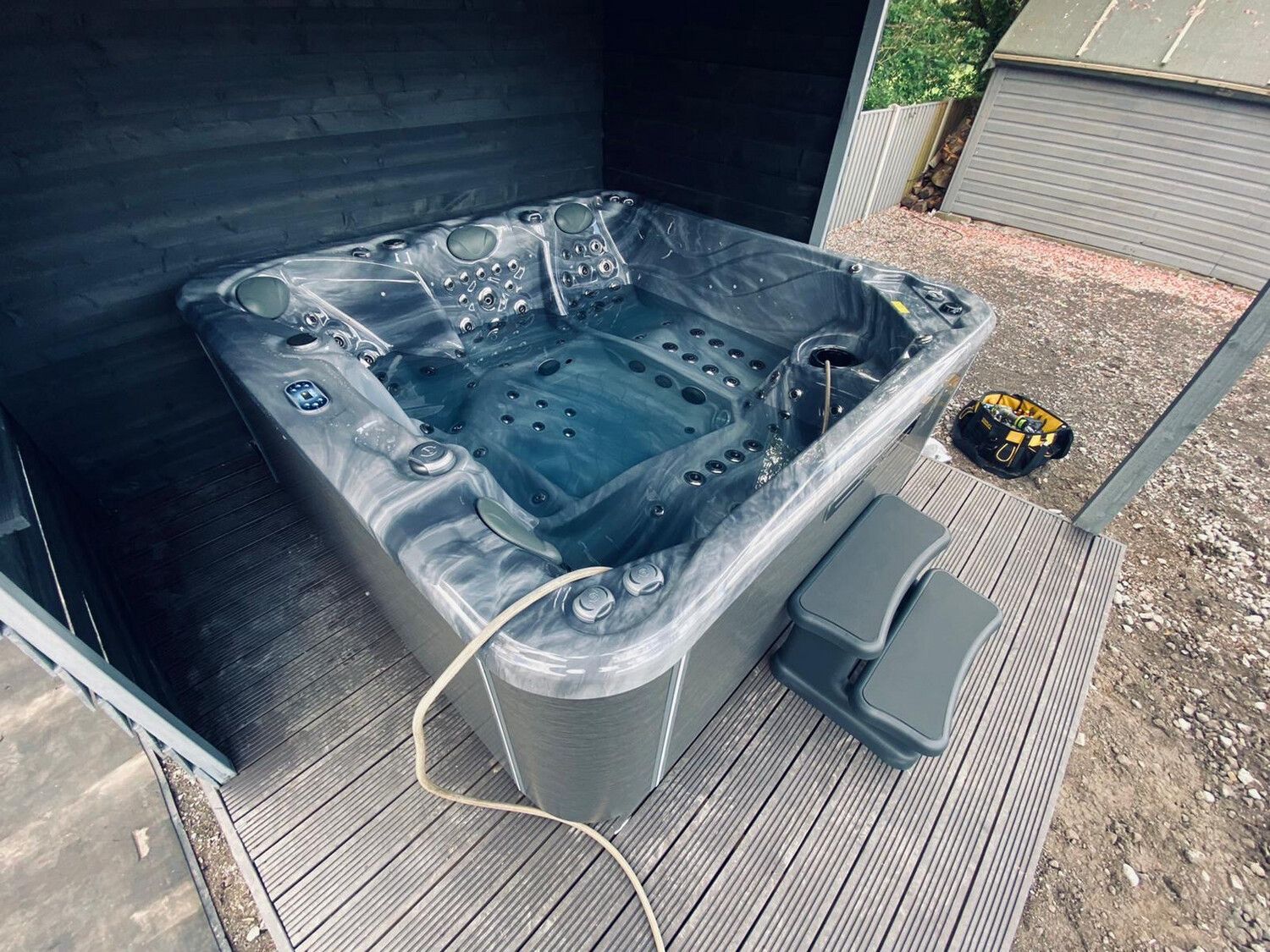 Gray hot tub with steps on a wood deck, enclosed by black walls.