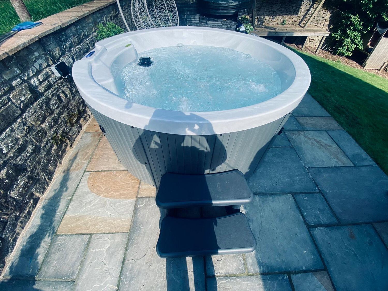 A round, gray hot tub with bubbling water sits on a stone patio, with steps leading up to it.