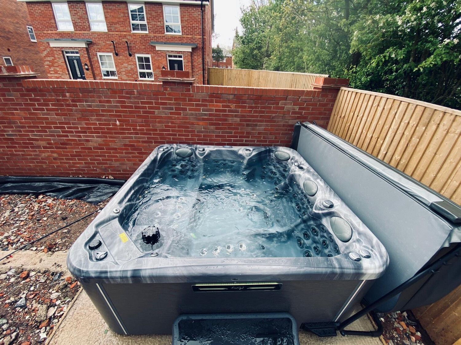 Hot tub with bubbling water sits against a brick wall and wooden fence outdoors.