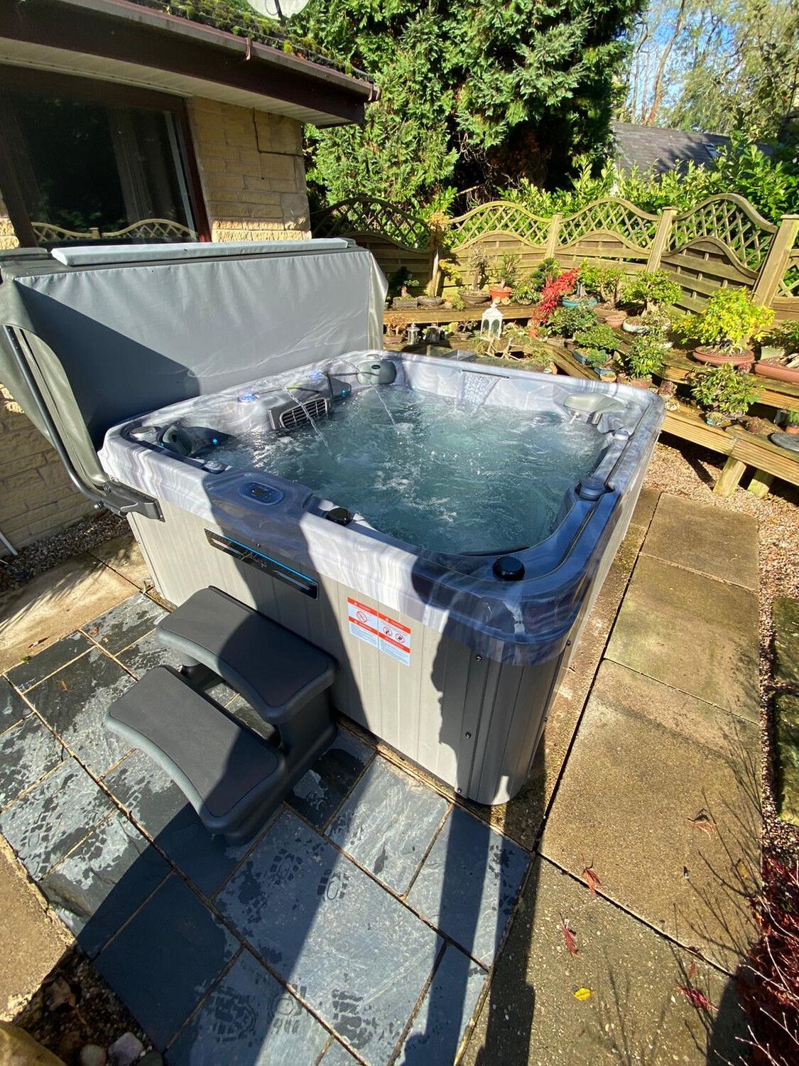 Hot tub filled with water sits on a patio. Steps lead up to the tub, with the cover open.