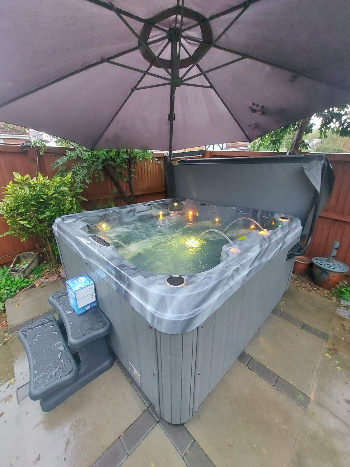 Hot tub with open cover under a gray umbrella, steps to the side, set in a backyard.