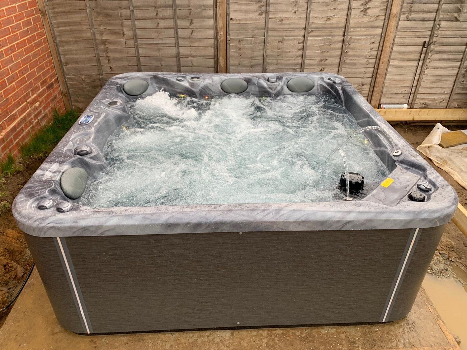 A dark gray hot tub with bubbling water sits outdoors near a fence.