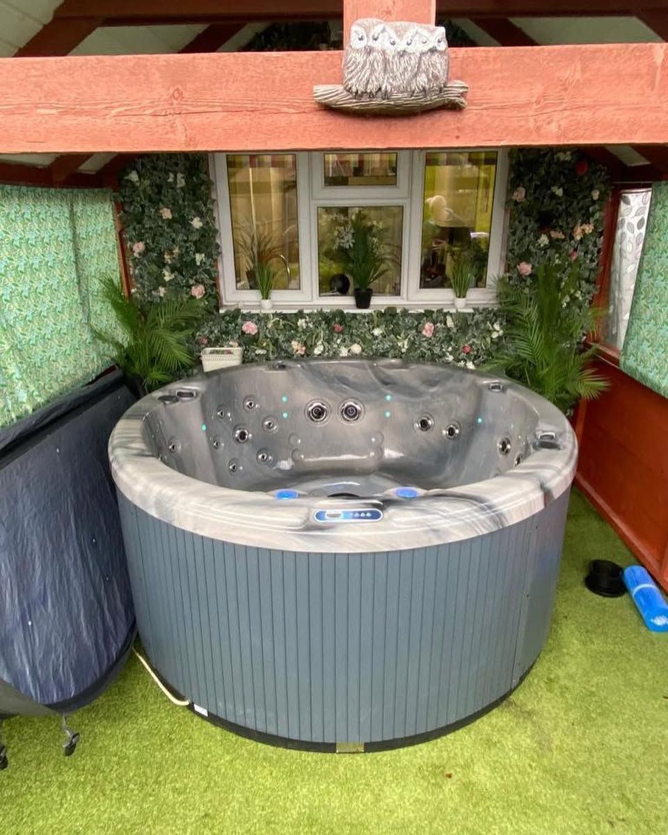 Round, blue hot tub in a covered outdoor space with a window and greenery.
