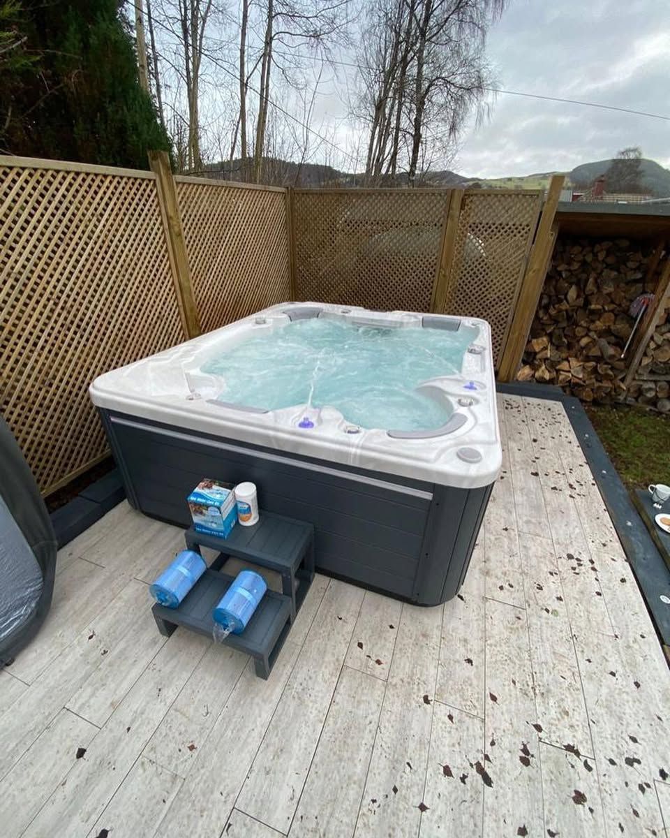 Hot tub with bubbling water on a tiled patio, next to a wooden fence and logs.