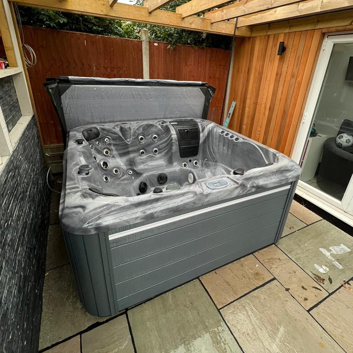 Hot tub with open cover, set on stone patio in a covered outdoor area with wooden siding and a sliding glass door.