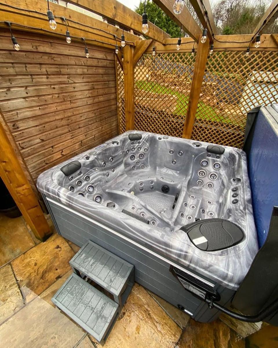 Hot tub on patio with steps, surrounded by wooden structure and greenery.