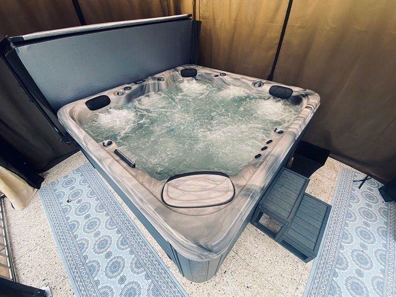 A bubbling, grey hot tub with cover open, on a speckled floor, and blue patterned rugs.