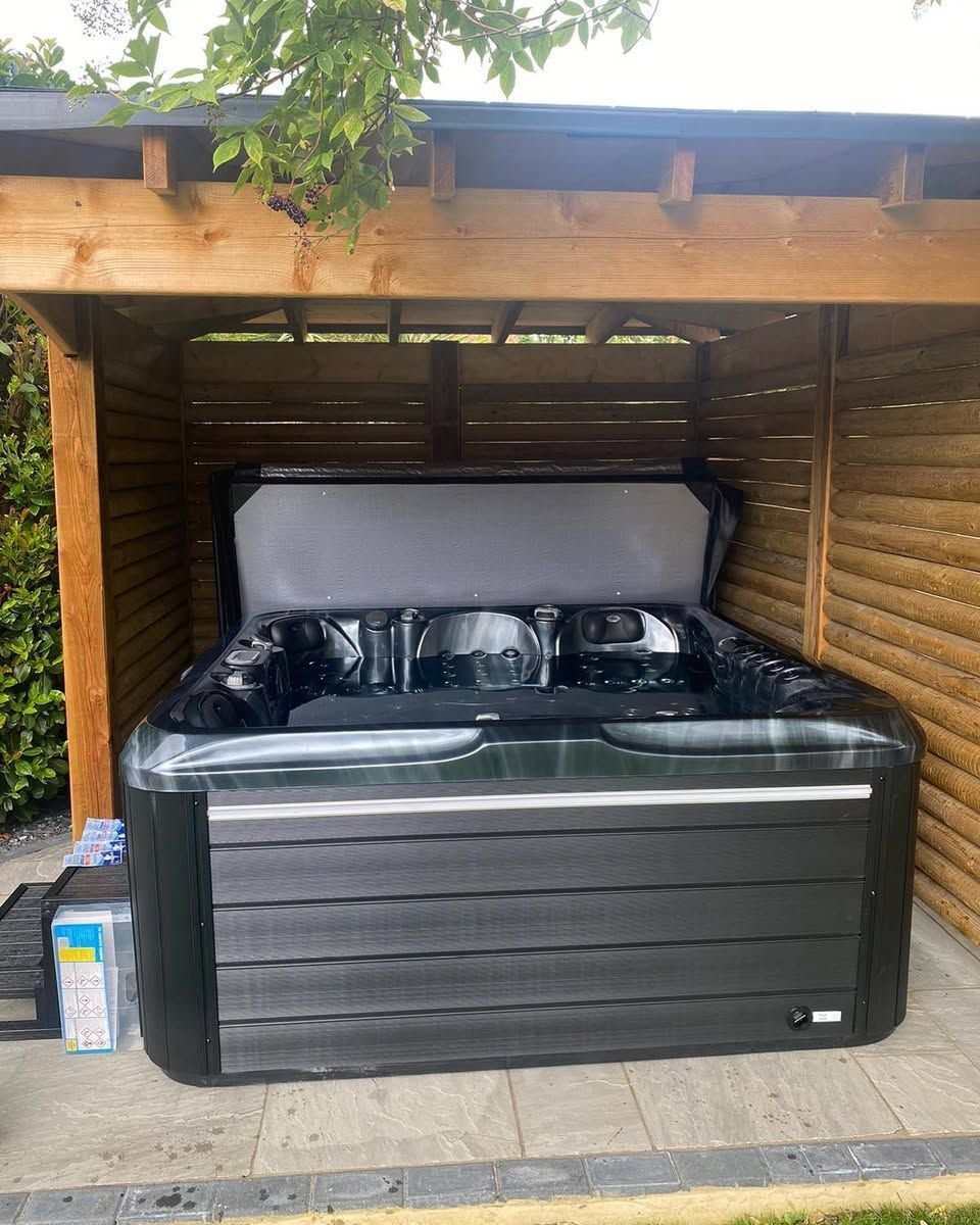 Black hot tub under a wooden pergola on a tiled patio. The lid is open.