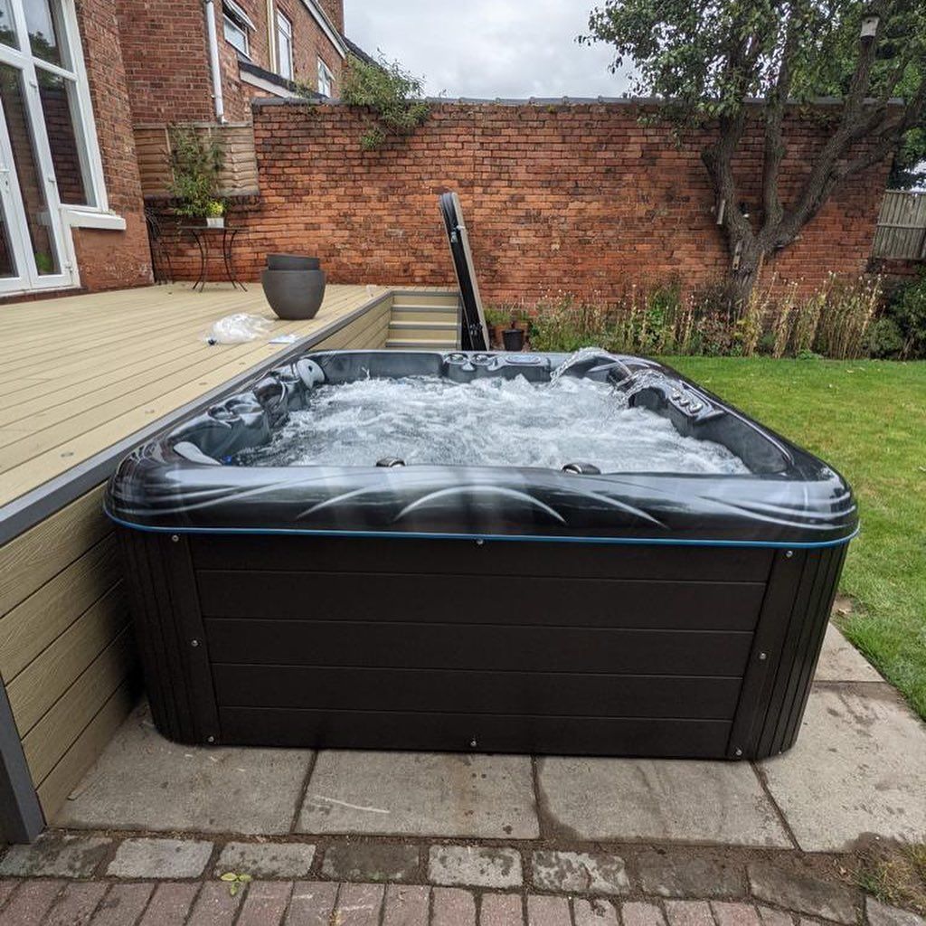 Hot tub in a backyard setting, next to a deck and brick wall. Black exterior, water bubbling.