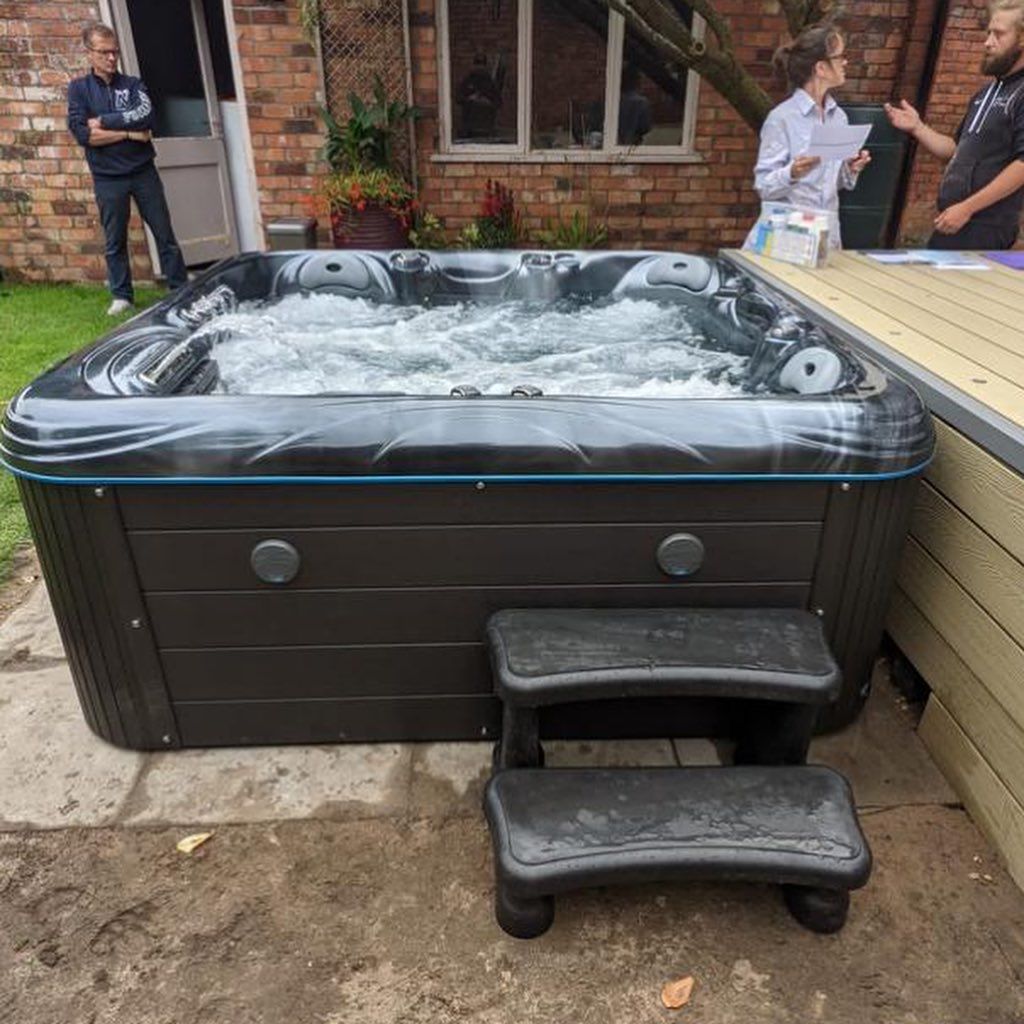 Hot tub with bubbling water and attached steps, in a backyard setting. People in the background.