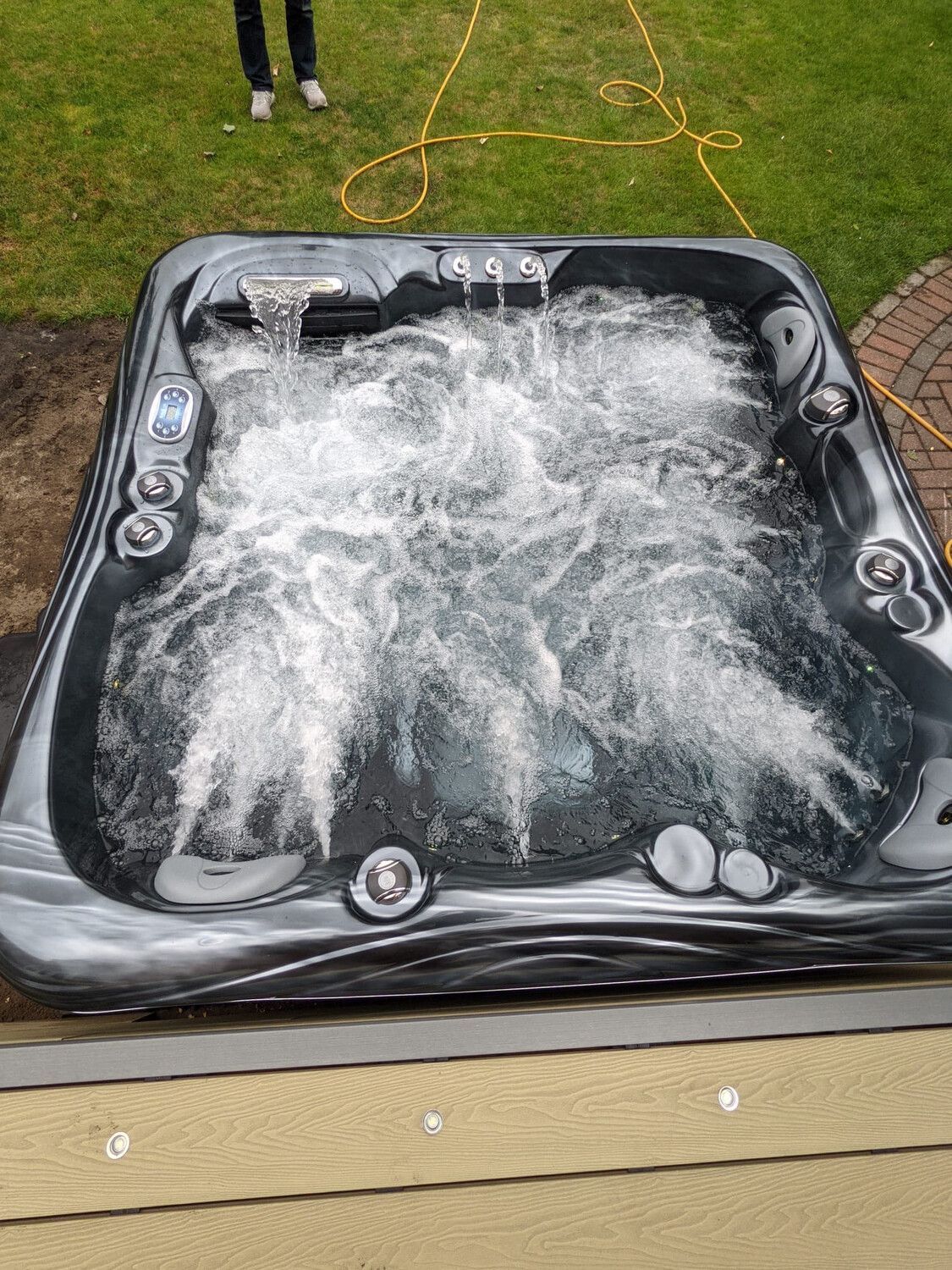 A hot tub with water jets activated, creating bubbles. Set outdoors on a deck, with a person standing nearby.