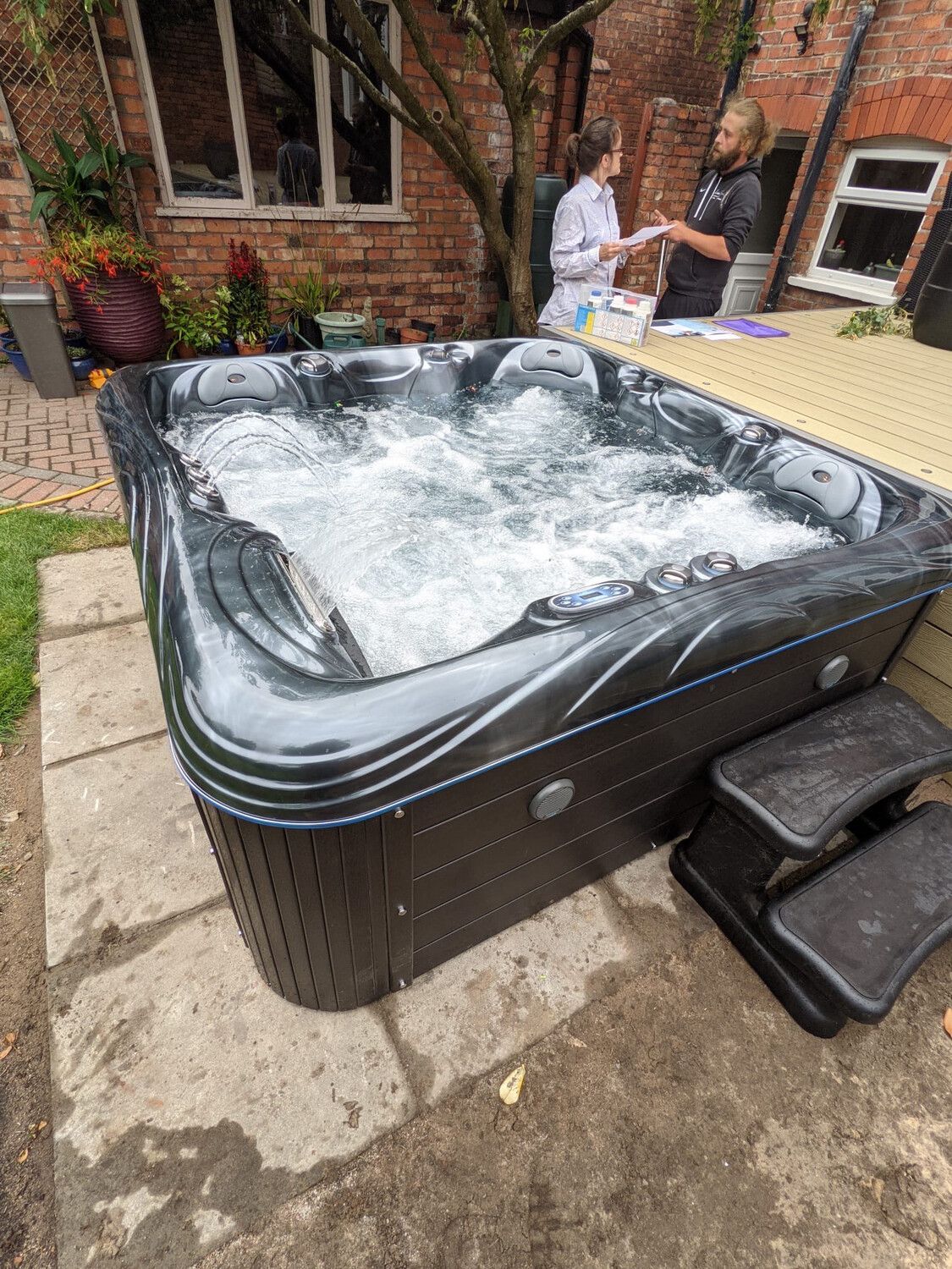 A bubbling black hot tub on a patio with two-step stairs; two people stand nearby.