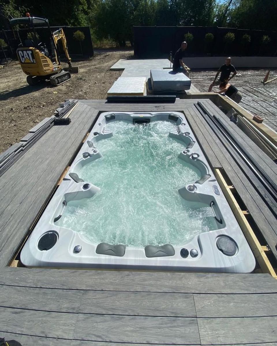 A rectangular hot tub is installed in a wooden deck. Construction site setting. Two men work nearby, small excavator.