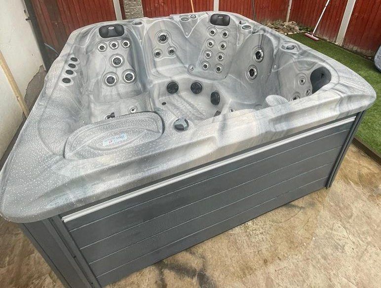 Gray hot tub with multiple jets, placed on a concrete patio next to some grass and a red fence.