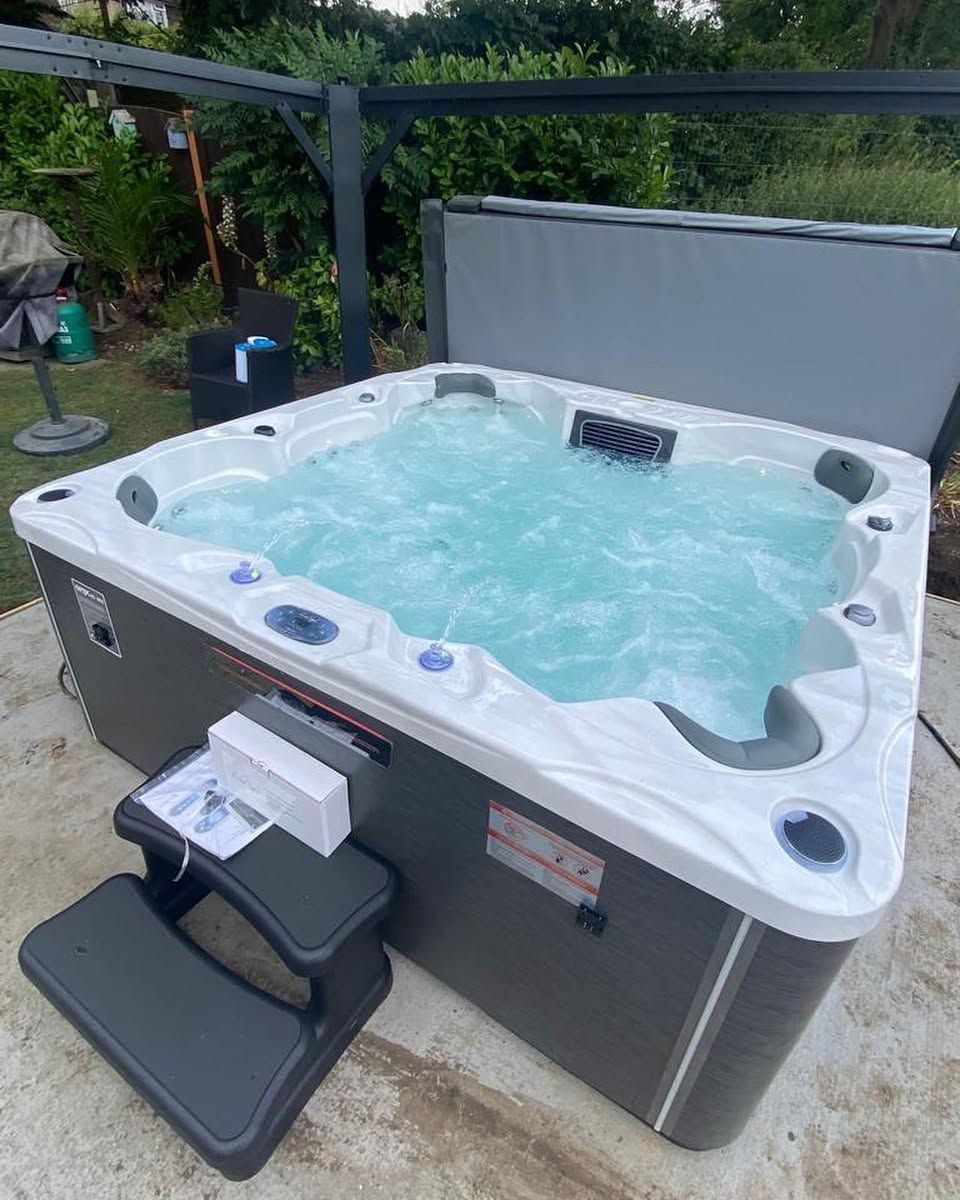 A hot tub filled with water, grey exterior, with steps on a concrete patio.