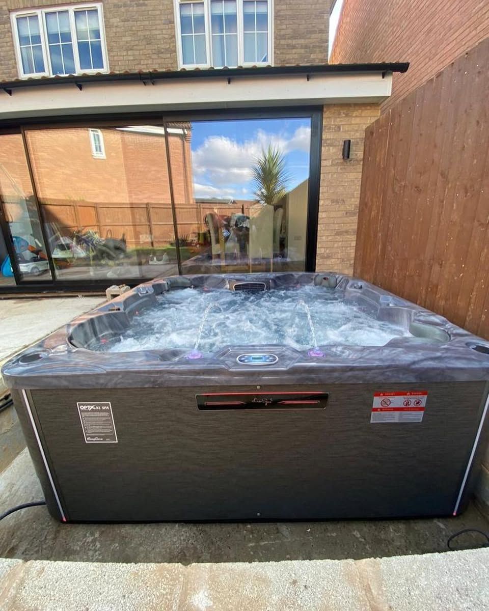 Hot tub with bubbling water sits on a concrete patio, reflecting the sky and surrounding house.