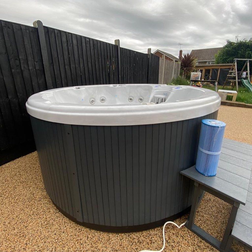Oval hot tub with gray exterior and white rim, near a black fence, on pebble ground, with filter and small table.