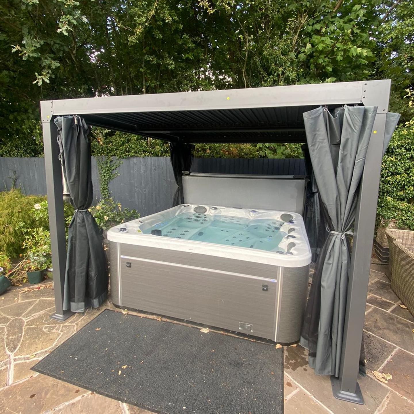 Hot tub under a gray pergola with dark curtains; stone patio and greenery in the background.