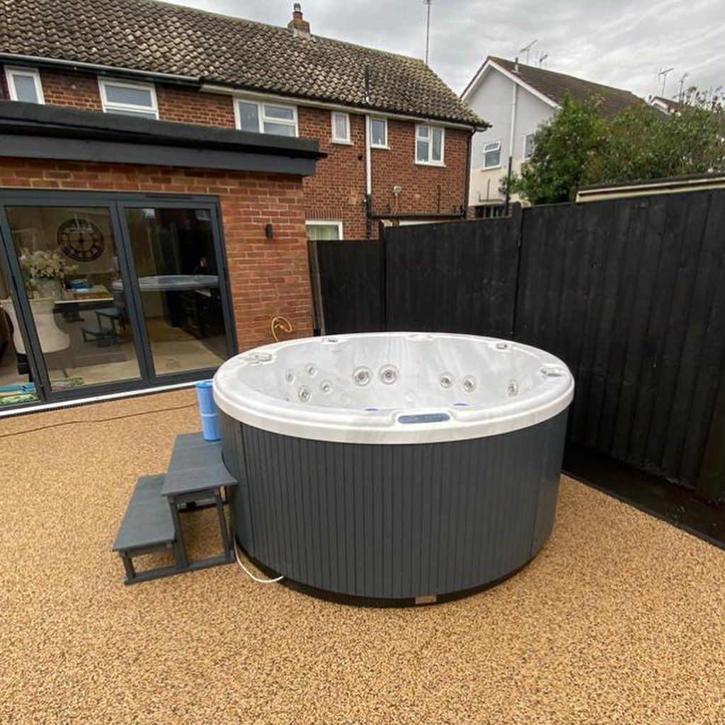 Hot tub on a speckled patio near a house and black wooden fence.