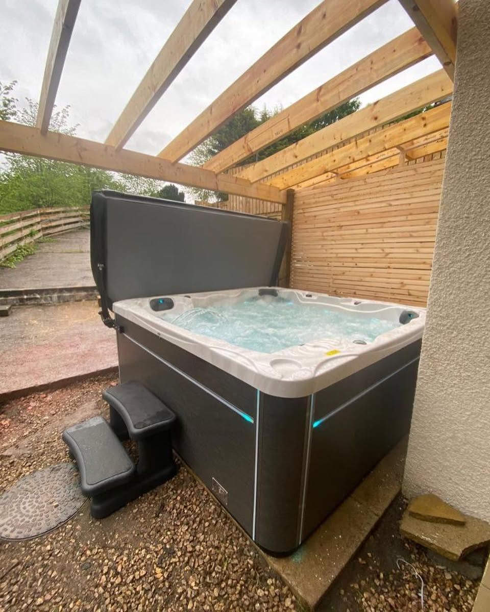 Hot tub with open cover, steps, under wooden pergola.  Dark gray and white, built-in side lighting, outdoors.