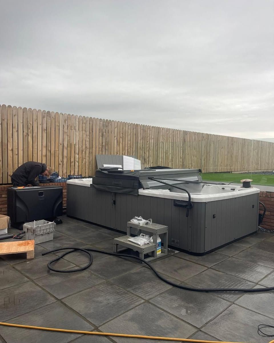 Hot tub being set up outdoors. Person near the black side unit. Gray tub on a tiled patio, brown fence in the background.