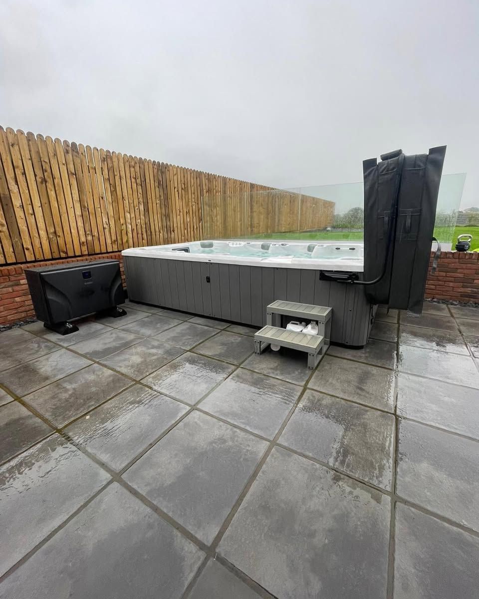 Outdoor hot tub on a tiled patio. Wooden fence and protective covers visible. Cloudy, overcast sky.