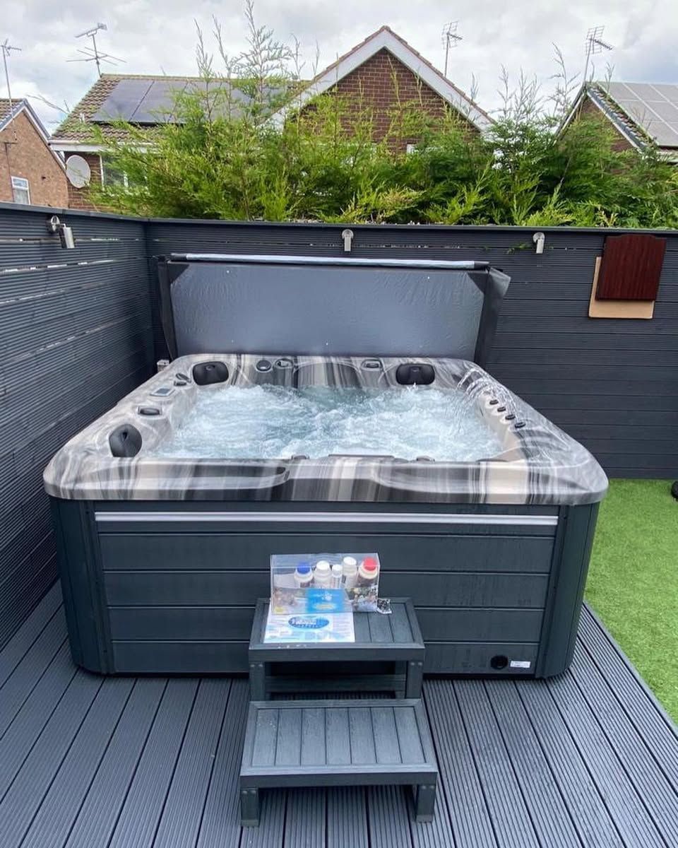 Hot tub on gray deck, cover open, bubbles visible. Step stool in front. Black privacy fence and greenery in background.