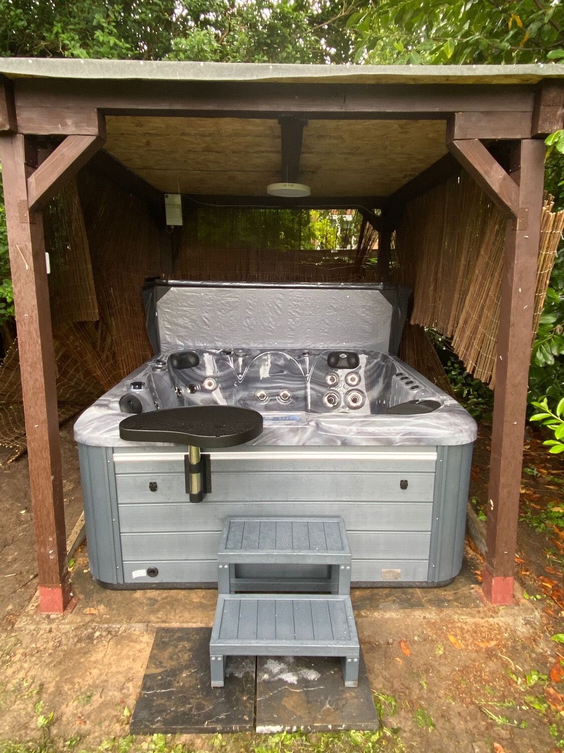 Hot tub under a wooden gazebo with steps, in an outdoor setting. Gray tub with gray steps.