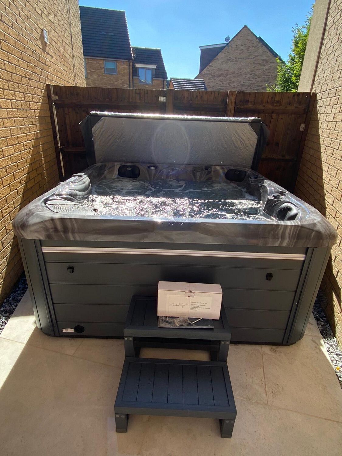 Hot tub with open cover, dark gray on a patio, small steps in front.