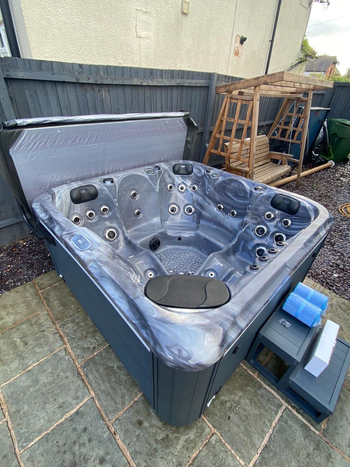 Dark blue hot tub with open cover, on a patio, near a wooden swing set and gray fence.