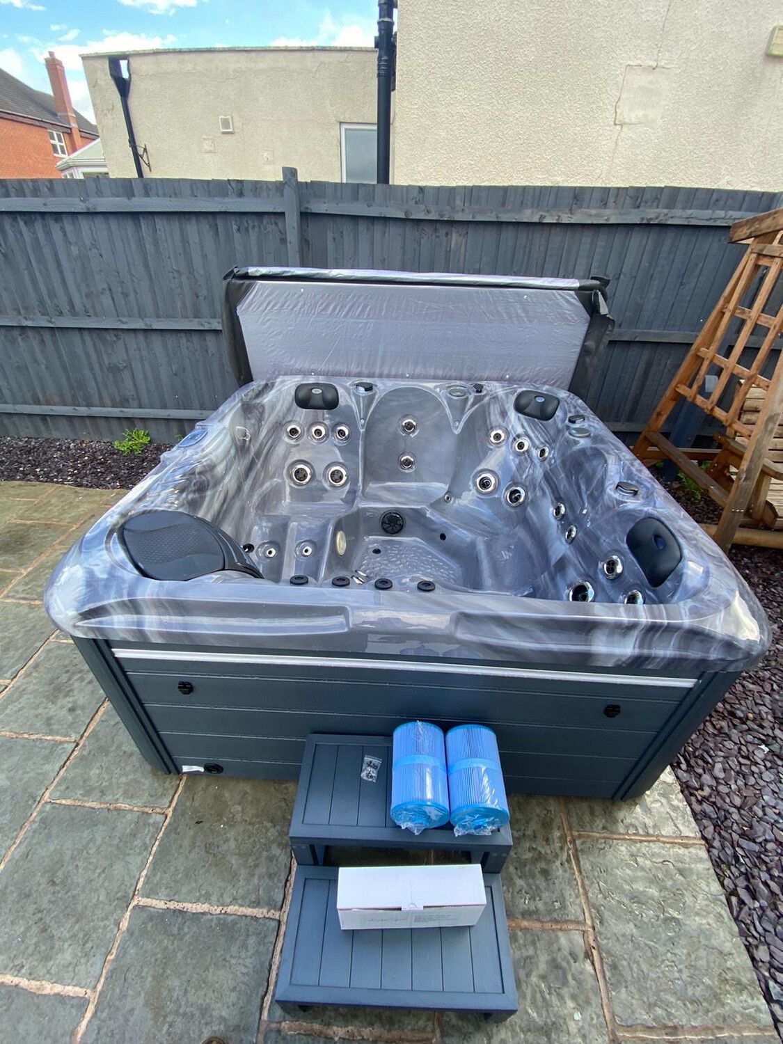 Dark gray hot tub with open cover, steps, filters, and chemicals in a backyard setting.