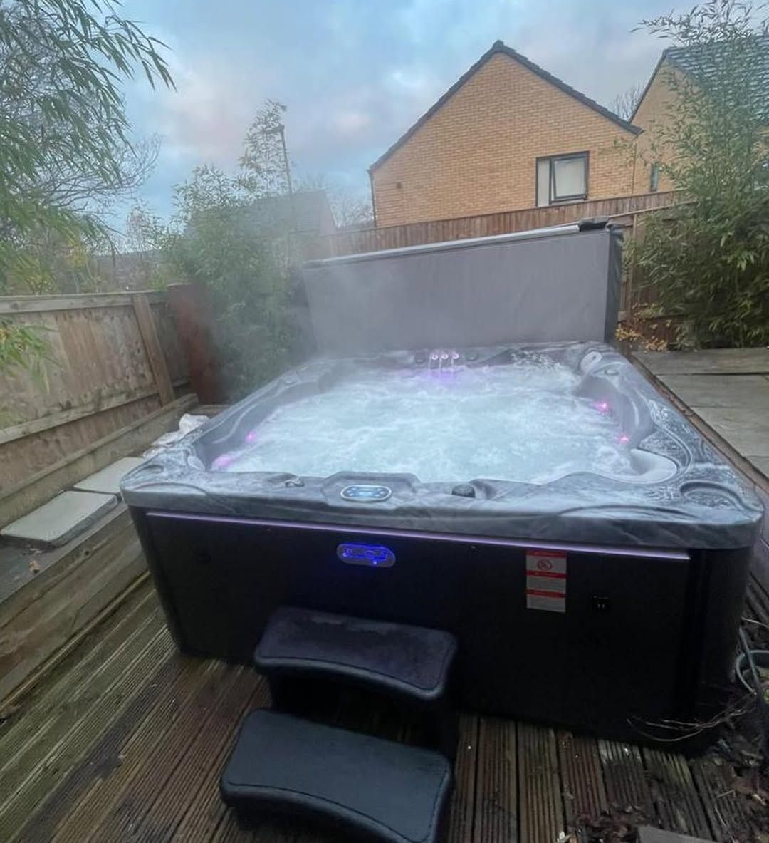 Hot tub with steam rising in a backyard setting. Dark tub with illuminated jets, surrounded by wooden deck and fence.