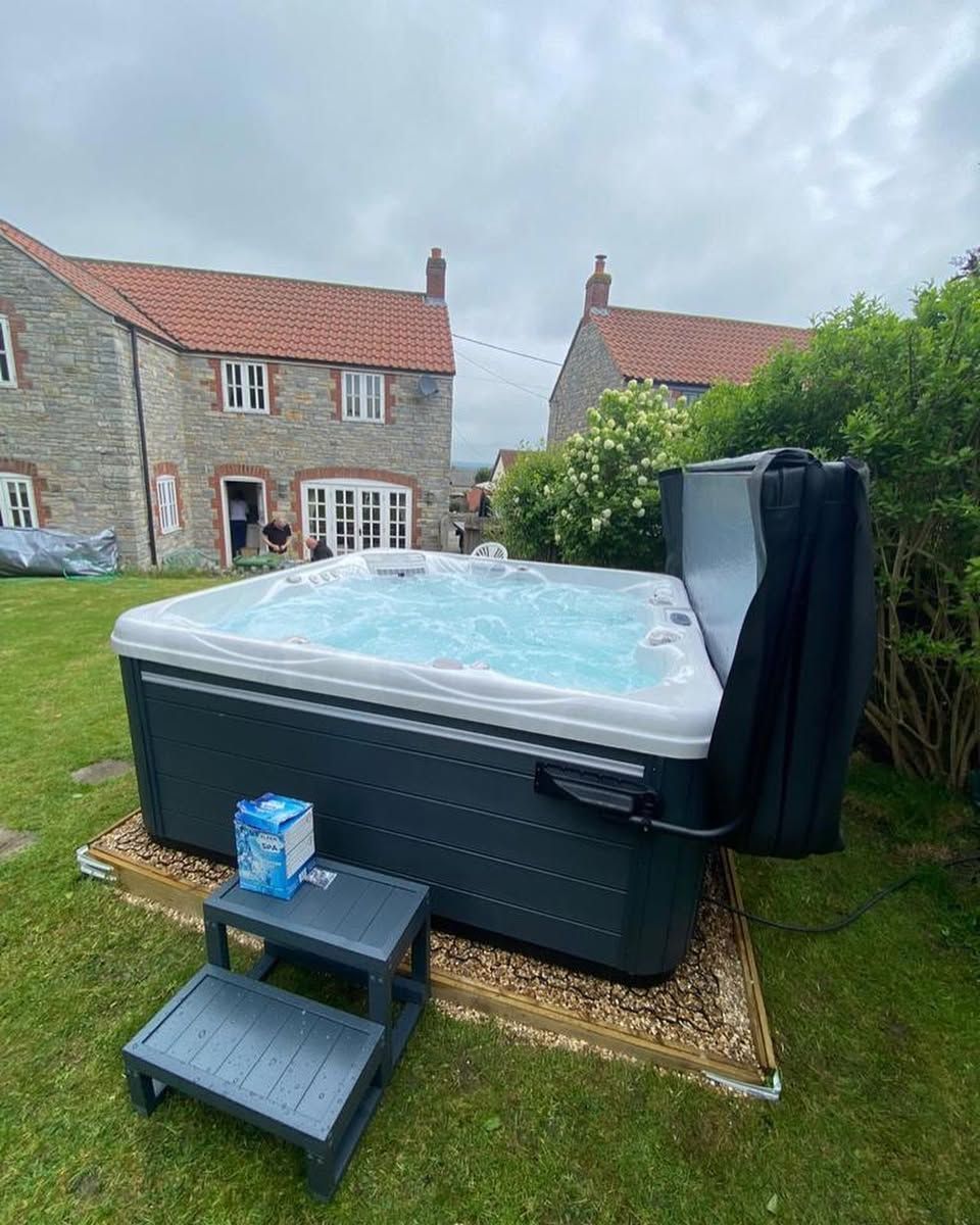 Hot tub on a lawn in front of stone houses with steps, cover, and water treatment supplies.