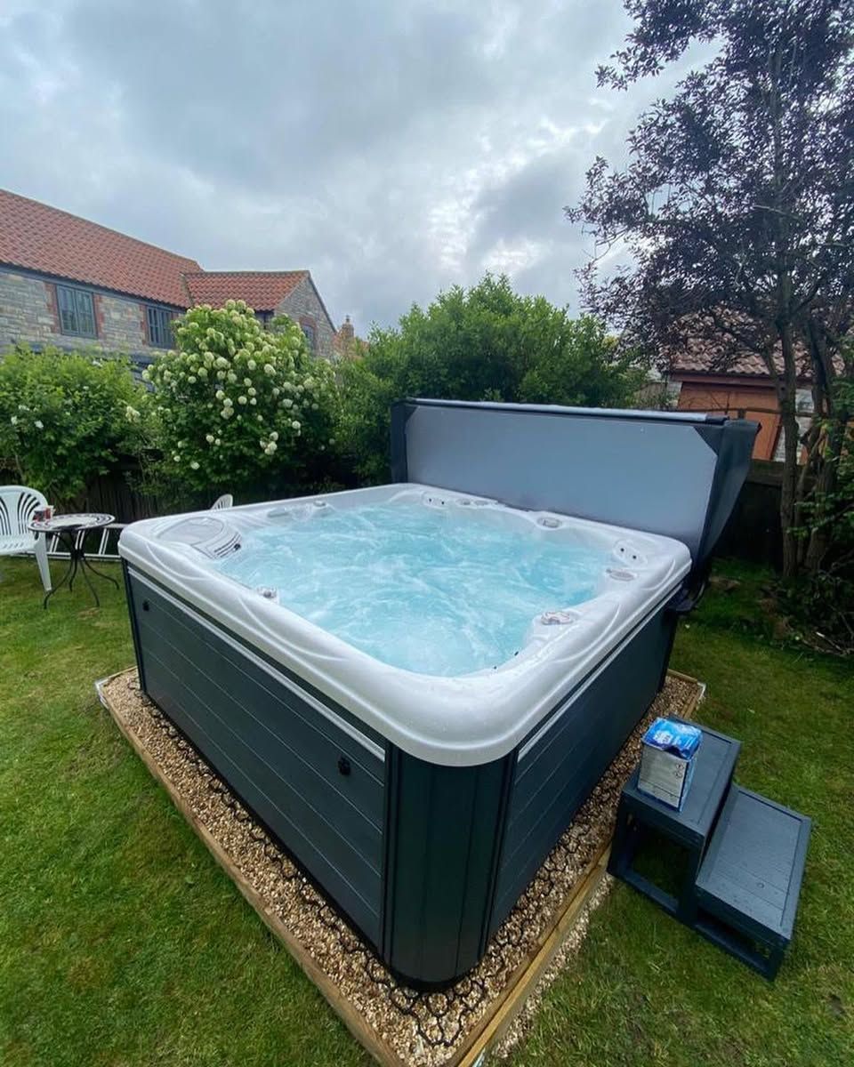 Hot tub with partially open cover in a grassy yard. Gray siding, blue water, and step for entry. Overcast sky.