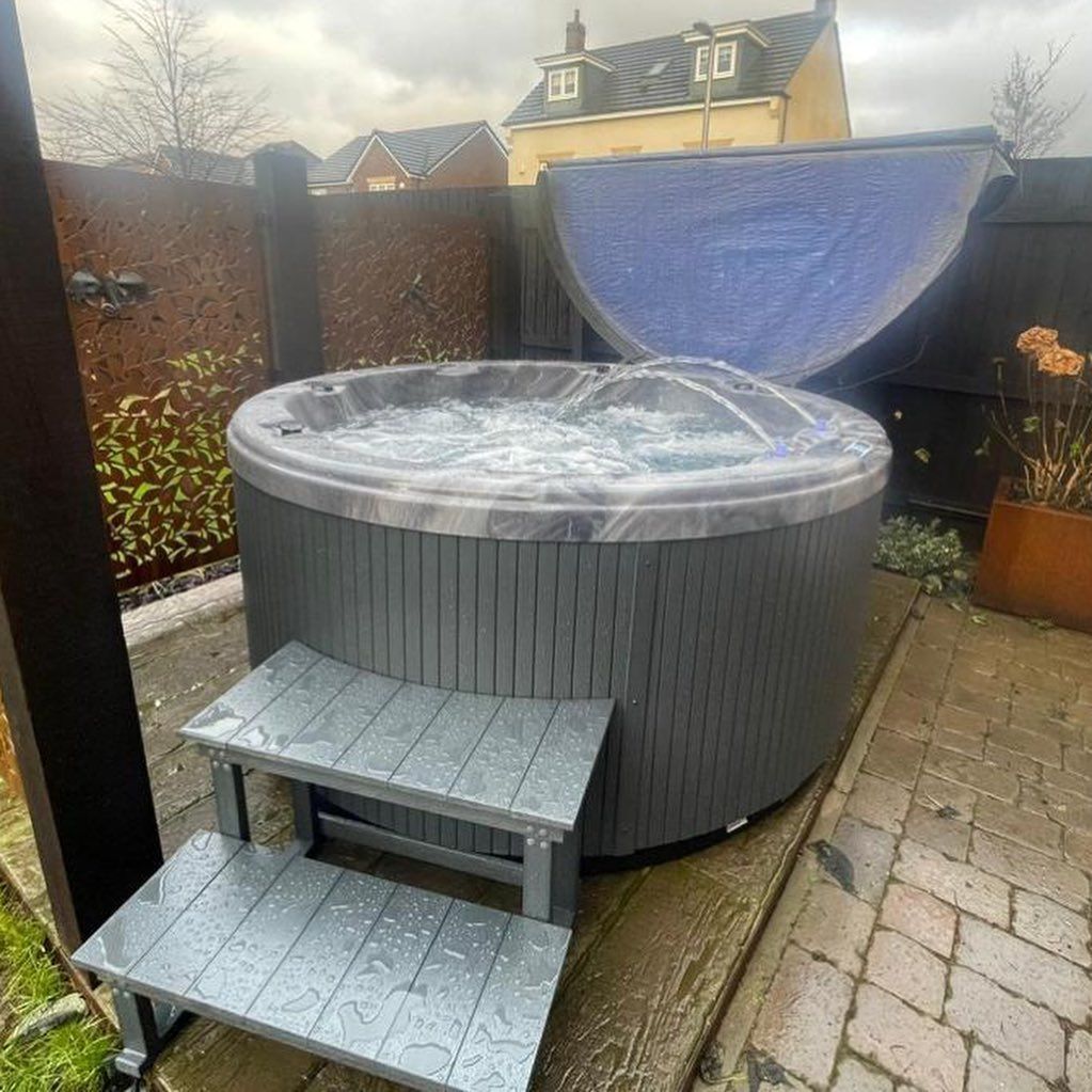 A gray hot tub with water, open blue cover, and steps in a backyard setting.