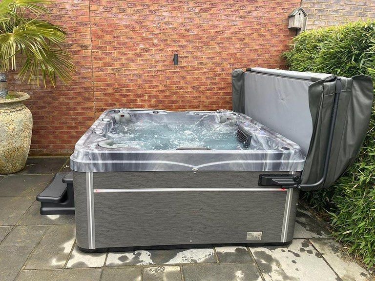 Hot tub with open cover, bubbling water, set on patio near a brick wall and foliage.