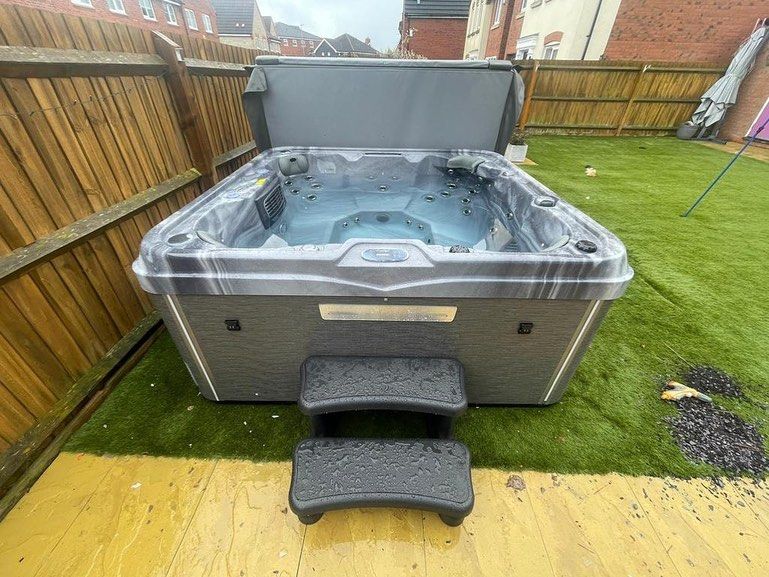Hot tub on a green lawn with open cover, steps in front. Grey and black. Wooden fence in background.