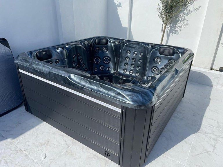 Dark gray square hot tub on a tiled patio, next to a white wall and an olive tree.