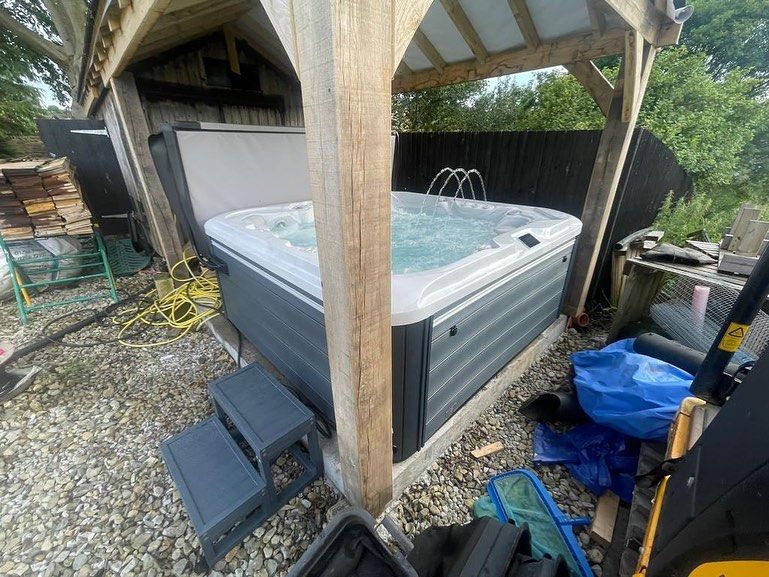 A hot tub under a wooden gazebo with steps, surrounded by yard tools and a fence.