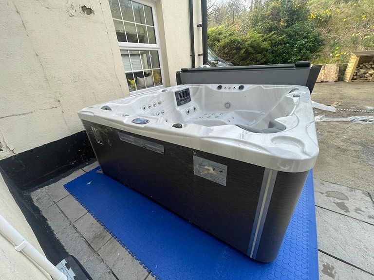 A dark-colored hot tub with a light-colored top sits outside near a window.