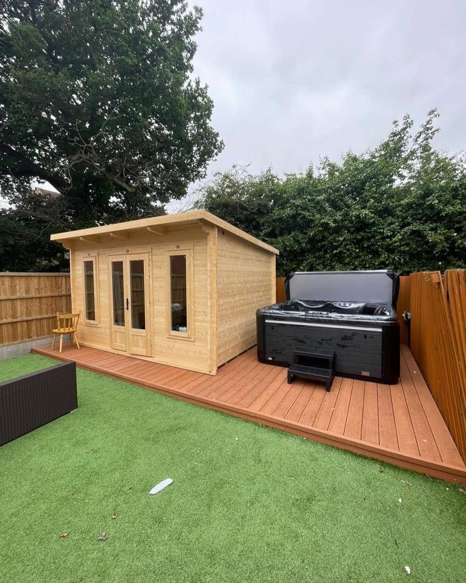 Wooden cabin and hot tub on a deck in a backyard with artificial turf and a wooden fence.