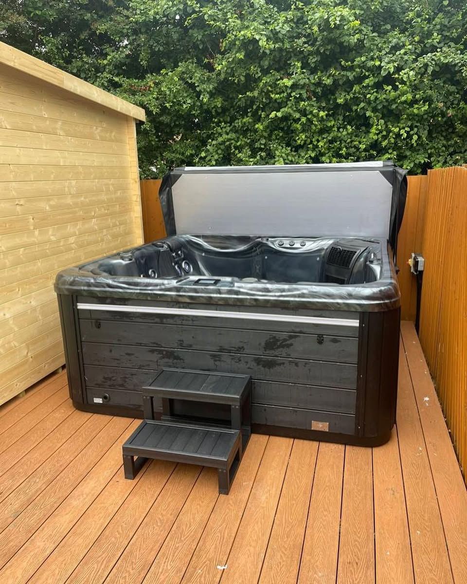 Hot tub with open cover, black exterior, wooden deck, small step stool, near a wooden shed.