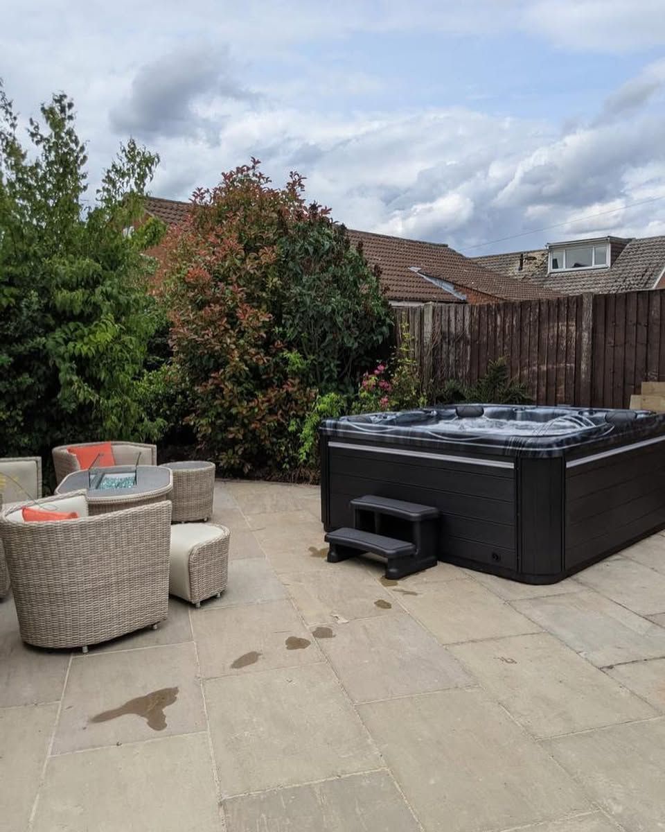 Patio with hot tub and outdoor seating against a backdrop of trees and a cloudy sky.