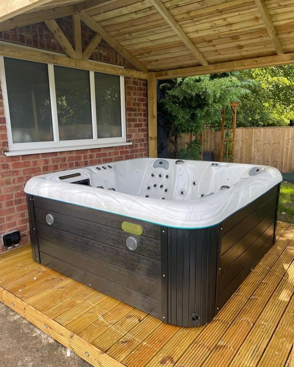 A hot tub under a wooden pergola on a wooden deck next to a brick building.