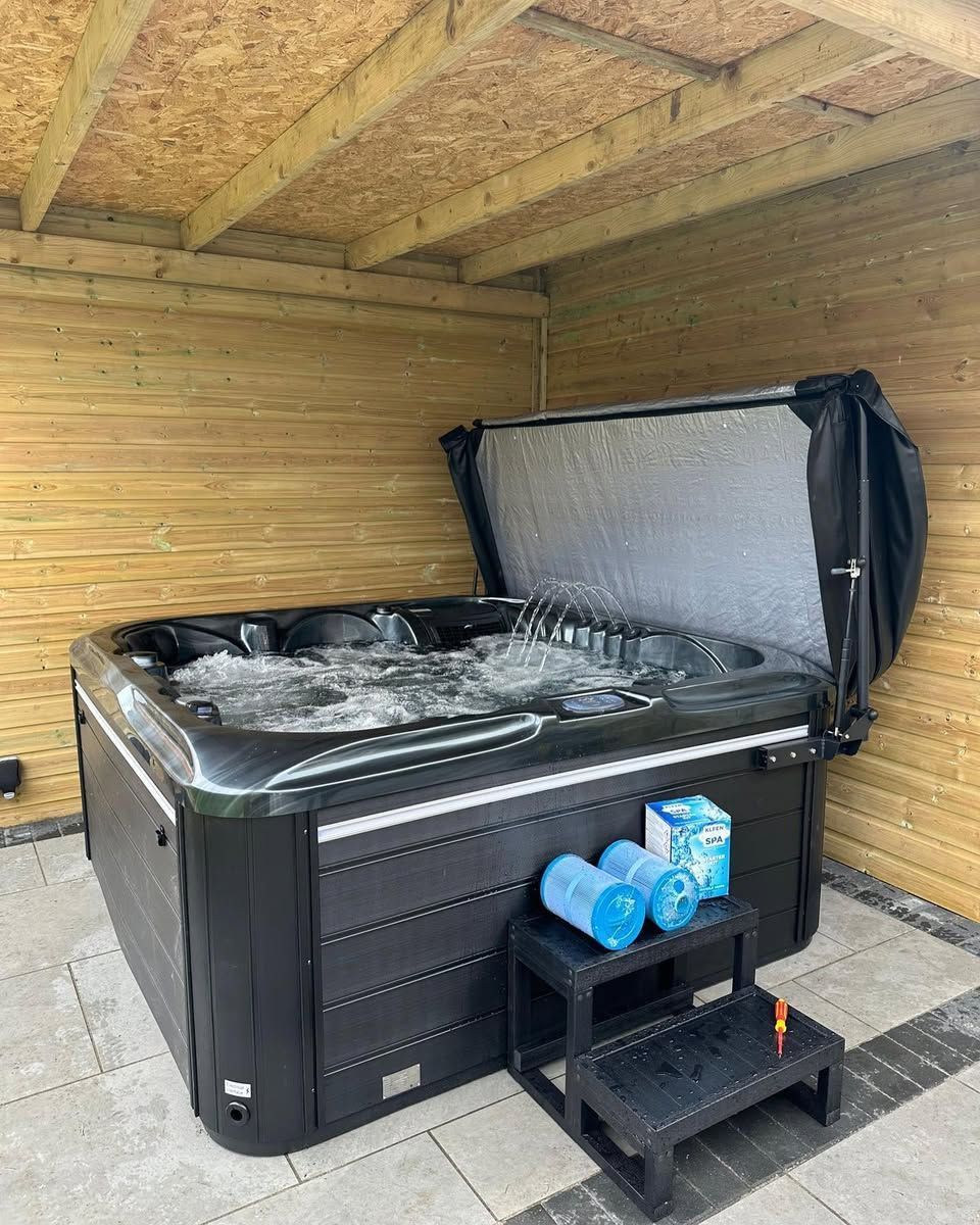 Hot tub with open cover, in a wooden enclosure. Black tub with jets, on a tiled patio.
