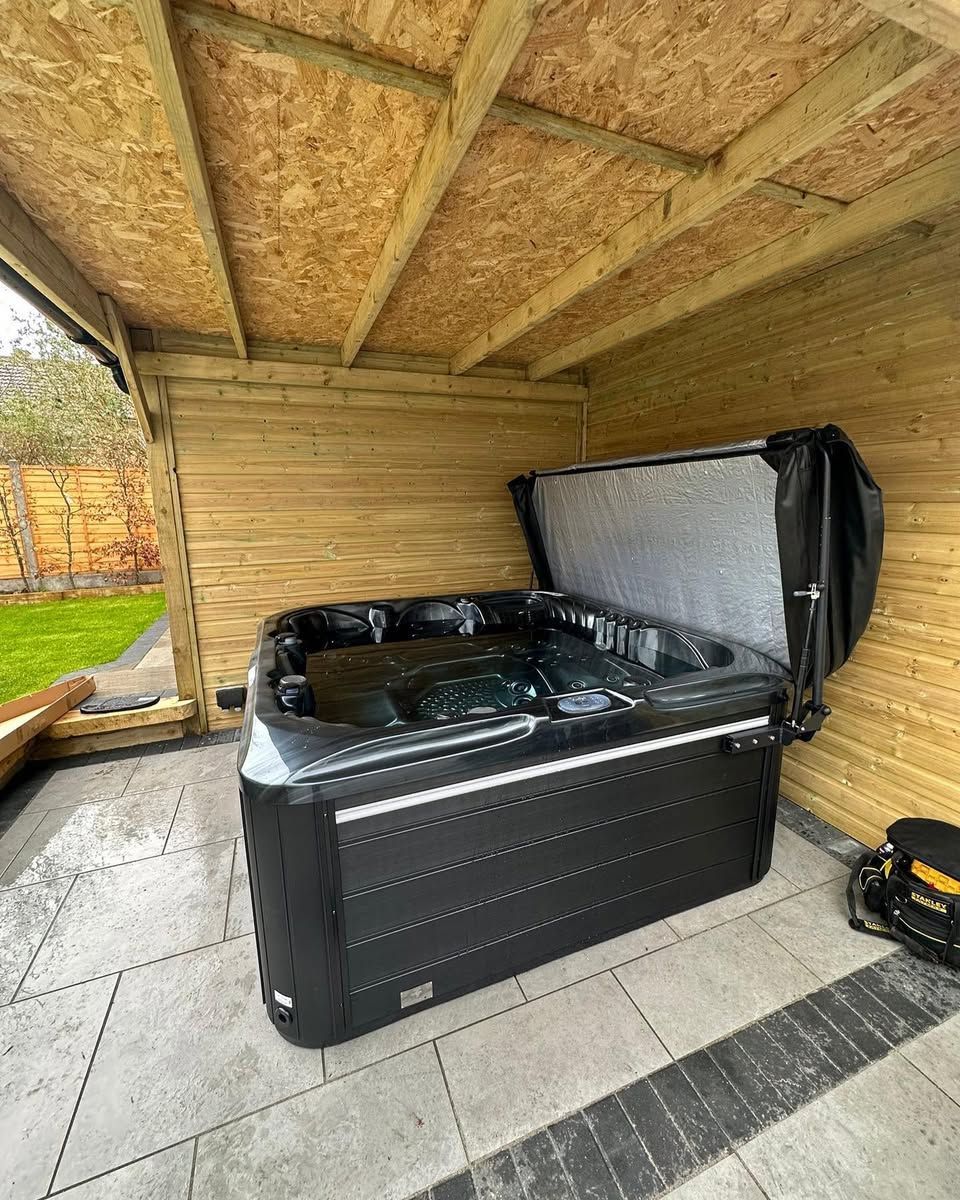 Black hot tub under a wooden shelter, cover open, on a tiled patio.