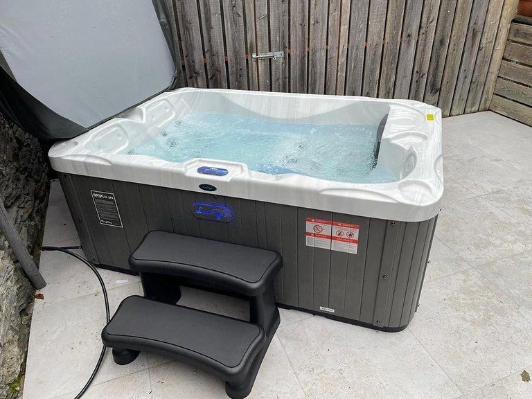 Hot tub filled with water and bubbles, next to two-step black stairs on a patio. Wooden fence in the background.