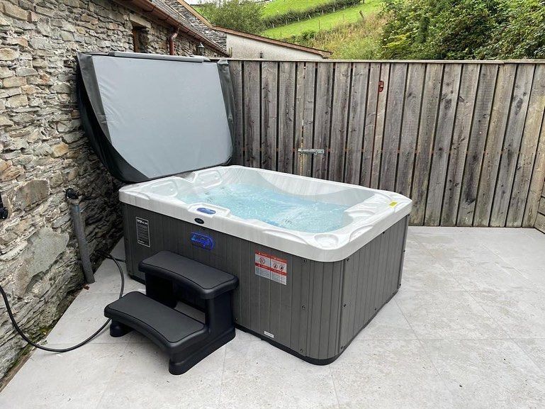 Hot tub with open lid, steps, on a patio next to a stone wall and wooden fence.